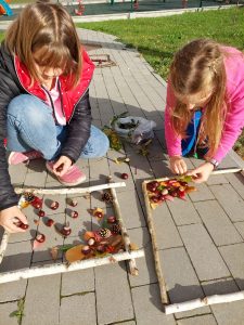 Dwie uczennice kucają na chodniku przy szkole i w ramkach ułożonych z patyków tworzą przy przy użyciu szyszek i orzechów prace.