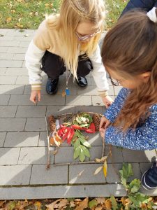 Dwie uczennice kucają na chodniku. Przed nimi ułożona jest ramka z patyków w której układają liście o szyszki tworząc jesienny obraz.
