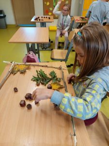 Uczennica klasy drugiej siedzi przy stoliku i tworzy obraz z darów jesieni. W ramce zbudowanej z patyków układa liście i kasztany.