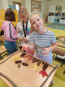 Uczennica stoi w sali lekcyjnej. Przed dziewczynką na stoliku ułożona jest ramka z patyków a w niej rozłożone liście, szyszki i kasztany.