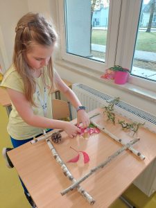 Uczennica stoi przy stoliku w sali. Przed nią na stole ułożona jest ramka z patyków, w której dziewczynka układa liście i szyszki tworząc jesienny obraz.