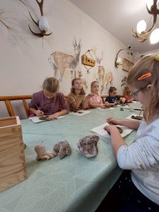 Uczniowie siedzą przy długim stole. W pomieszczeniu na ścianach widoczne są rysunki zwierząt leśnych. Stół zakryty jest zielonym obrusem, a na nim uczniowie formują glinę.