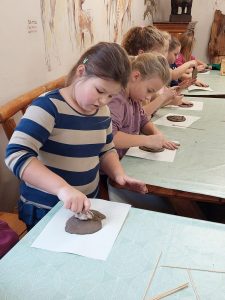 Uczniowie siedzą przy długim stole zakrytym zielonymi obrusami. Dzieci odciskają w glinie tropy zwierząt.