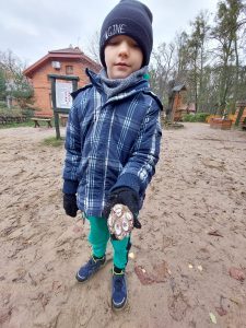 Uczeń stoi na ziemi. W tle widać zabudowania leśnej szkoły na Barbarce. Chłopiec w wyciągniętej dłoni trzyma odlew odcisku leśnego zwierzęcia.