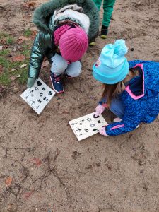 Uczniowie pochylają się nad ziemią w poszukiwaniu tropów zwierząt. Dwie uczennice widoczne na zdjęciu mają przed sobą kartki, na których odciśnięte są i podpisane tropy różnych zwierząt.