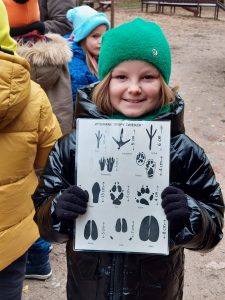 Uczennica prezentuje trzymając przed sobą kartę z tropami zwierząt. Każdy odcisk jest podpisany i zwymiarowany.