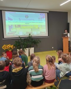 Uczniowie siedzą na ławeczkach przed rzutnikiem na którym wyświetlone jest pytanie z quizu o Janie Pawle II.