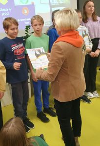 Pani Dyrektor wręcza uczniowi stojącemu na przeciwko dyplom. Obok w rzędzie stoją inny uczniowie.