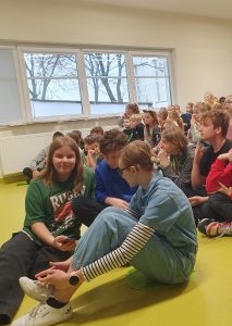 Uczniowie siedzą na podłodze. Trójka uczniów siedząca na pierwszym planie spogląda w telefon trzymany przez jedną z uczennic.