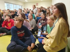 Uczniowie na pierwszym planie siedzą na podłodze w sali teatralnej. Patrzą w lewą stronę. Jeden z chłopców trzyma telefon. Za nimi widać siedzących na krzesełkach innych uczniów.