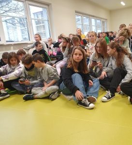 Uczniowie siedzą na podłodze w sali teatralnej. Widoczne są dwie trzyosobowe grupki, które wspólnie rozwiązują zadania na telefonie.