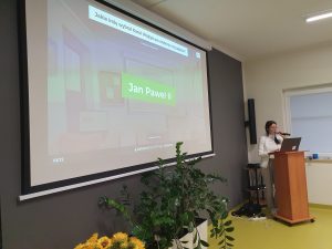 Nauczycielka religii stoi przy mównicy z mikrofonem w dłoni. Obok niej na rzutniku wyświetla się na zielonym tle napis "Jan Paweł II".