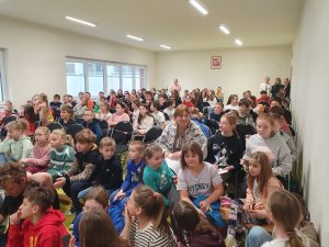 Uczniowie szkoły siedzą na krzesełkach w sali teatralnej i obserwują rozgrywki między drużynami.