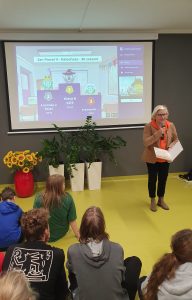 Pani Dyrektor stoi przed uczniami z mikrofonem w dłoni i otwartą sztywną podkładką. W tle na rzutniku wyświetla się ranking uczniów. Na pierwszym miejscu Klasa 6 KPZ na drugim Ananasy z klasy a na trzecim Kremówki.