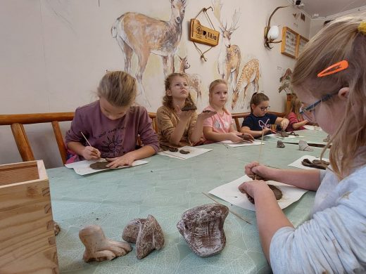 Uczniowie siedzą przy długim stole. W pomieszczeniu na ścianach widoczne są rysunki zwierząt leśnych. Stół zakryty jest zielonym obrusem, a na nim uczniowie formują glinę.