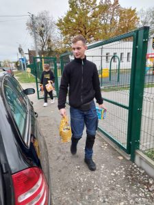 Pracownik szkoły idzie obok bramy niosąc w dłoniach torby z karmą dla zwierząt.