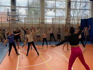 Uczennice uczestniczące w zajęciach z zumby stoją na przeciw trenerki i wymachują na boki dłoniami.