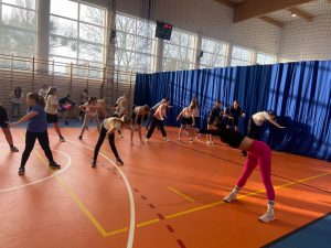 Uczennice uczestniczące w zajęciach z zumby stoją na przeciwko trenerki i robią skłony w przód wyciągając przed siebie jedną z rąk.