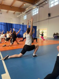 Uczennice uczestniczące w zajęciach z zumby klęczą na jednym kolanie z wyciągniętą w tył jedną nogą. Ręce wyciągają w górę