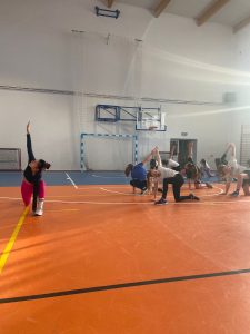 Uczennice uczestniczące w zajęciach z zumby powtarzają ćwiczenie prezentowane przez trenerkę. Klęcząc na jednej nodze z wyprostowaną drugą wysoko wyciągają jedną dłoń w górę a drugą opierają o podłoże.