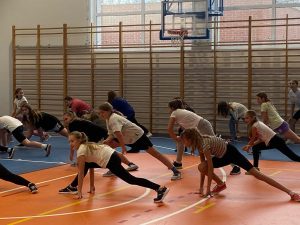 Uczennice uczestniczące w zajęciach z zumby robią szeroki rozkrok bokiem i opierają dłonie o podłogę przy zgiętym kolanie.