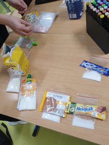 Na stole leżą poustawiane produkty wraz z odmierzonym cukrem pokazującym jego zawartość w poszczególnych produktach.
