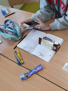 Uczennica klasy szóstej trzyma w dłoni telefon na którym przelicza zawartość cukru w produktach spożywczych. Na stole przed nią leży zeszyt, piórnik oraz opakowania po słodkich przekąskach.