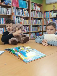Dwoje uczniów klasy pierwszej siedzi za dużym stołem w gminnej bibliotece w Ostaszewie. Dzieci przytulają misie. Na stole leży otwarta książka.