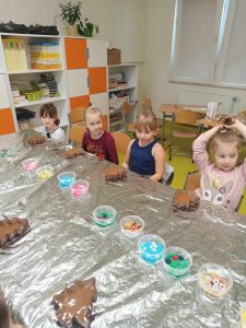 Uczniowie klasy pierwszej siedzą przy długim stole zakrytym folią aluminiową. Na stole ustawione są kubeczki z kolorowymi ozdobami do czekolady. Przed dziećmi leżą foremki w kształcie choinek wypełnione czekoladą.