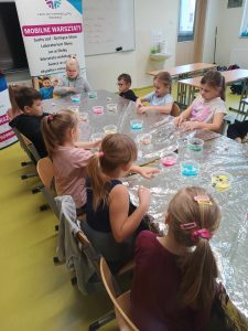 Uczniowie klasy pierwszej siedzą przy długim stole zakrytym folią aluminiową. Na stole ustawione są kubeczki z kolorowymi ozdobami do czekolady.