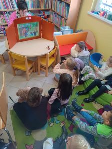 Uczniowie klasy pierwszej siedzą na dywanie w gminnej bibliotece w Ostaszewie. Dzieci oglądają teatrzyk przygotowany przez Panią bibliotekarkę częściowo ukrytą za pomarańczowym tryptykiem imitującym scenę teatru.