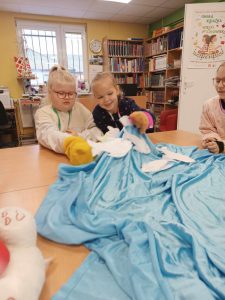 Dwie uczennice klasy pierwszej siedzą za dużym stołem w gminnej bibliotece w Ostaszewie. Przed nimi leży rozłożony niebieski materiał, a na nim wycięte z papieru rybki. Dziewczynki lekko podnoszą materiał do góry.