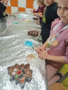 Uczniowie klasy pierwszej stoją przy stole zakrytym folią aluminiową. Przed dziećmi leżą foremki w kształcie choinek z przygotowanymi i ozdobionymi przez uczniów czekoladkami.