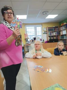 Dwie uczennice klasy pierwszej siedzą za dużym stołem w gminnej bibliotece w Ostaszewie. Przed nimi leżą misie. Jedna z nich spogląda na stojącą obok Panią bibliotekarkę, która trzyma w dłoni wyciętą z papieru i zalaminowaną grupę bohaterów serialu Gumisie.