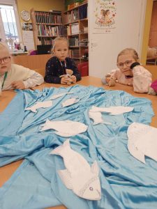 Trzy uczennice siedzą przy dużym stole w gminnej bibliotece w Ostaszewie. Dziewczynki spoglądają na rozłożony na stole niebieski materiał, na którym leżą wycięte z papieru ryby.