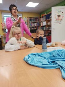 Dwoje uczniów klasy pierwszej siedzi za dużym stołem w gminnej bibliotece w Ostaszewie. Za nimi stoi Pani bibliotekarka z wyciągniętą w przód dłonią. Na stole leży niebieski materiał oraz szpula z nicią w kształcie stożka.