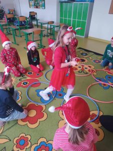 Uczennica klasy pierwszej przeskakuje przez rozłożone na dywanie hula hopy. Wokół niej na podłodze siedzą inni uczniowie w czapeczkach mikołajkowych.