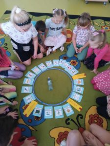 Uczniowie klasy pierwszej siedzą na podłodze w kręgu. Po środku leżą ułożone również po okręgu karteczki z różnymi napisami a wewnątrz tego kręgu leży butelka.