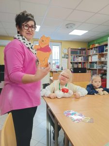 Dwie uczennice klasy pierwszej siedzą za dużym stołem w gminnej bibliotece w Ostaszewie. Przed nimi leżą misie. Jedna z nich spogląda na stojącą obok Panią bibliotekarkę, która trzyma w dłoni wyciętą z papieru i zalaminowaną postać Kubusia Puchatka.