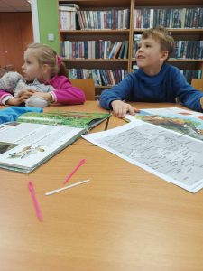 Dwoje uczniów klasy pierwszej siedzi za dużym stołem w gminnej bibliotece w Ostaszewie. Na stole leżą otwarte książki oraz kolorowe wąskie zakładki.