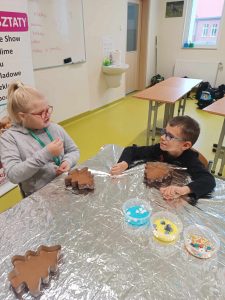 Dwoje uczniów klasy pierwszej siedzi przy stole zakrytym folią aluminiową. Przed nimi na stole leżą formki w kształcie choinek wypełnione czekoladą. Pośrodku stołu ustawione są trzy kubeczki z kolorowymi ozdobami do czekoladek.