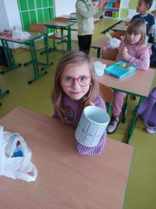Uczennica klasy pierwszej siedzi przy stoliku i wyciąga w dłoni przed sobą otrzymany kubek.