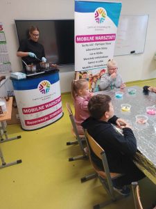 Na zdjęciu widoczny jest kontuar, za którym stoi Pani mieszająca czekoladę w garnach oraz część stołu zakrytego folią aluminiową. Za stołem siedzą uczniowie klasy pierwszej, a na stole porozstawiane są kubeczki z kolorowymi ozdobami do czekoladek.