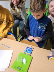 Uczniowie klasy czwartej stoją przy stole, na którym ustawiona jest waga oraz opakowanie po soczku ze słomką. Na wadze leży woreczek z cukrem, a uczniowie przyglądają się wskazaniom wagi.