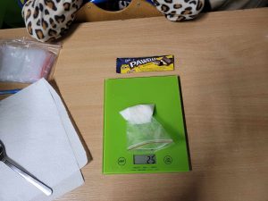 Na stole leży waga kuchenna na której ułożony został woreczek z cukrem. Waga wskazuje 25 g. Przed wagą leży opakowanie po batonie "PAWEŁEK".