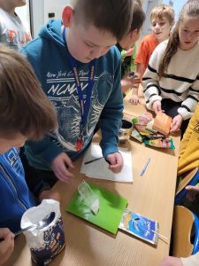 Uczniowie klasy czwartej stoją przy stoliku na którym ustawiona jest waga kuchenna. Na wadze ważony jest woreczek z cukrem. Przy wadze leży opakowanie po soczku ze słomką.