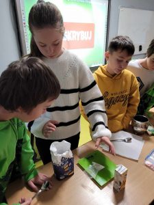 Uczniowie klasy czwartej ważą woreczek z cukrem na wadze kuchennej. Na stole leży obok wagi paczka cukru oraz opakowanie po soczku w kartoniku.