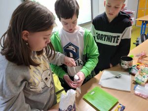 Uczniowie klasy czwartej odmierzają do strunowego woreczka cukier. Jedna z uczennic trzyma łyżeczkę z cukrem, a chłopiec stojący obok łyżeczkę.