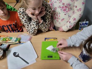 Uczniowie klasy czwartej przyglądają się wadze, na której ułożony jest woreczek z cukrem, do którego jedna z uczennic wkłada łyżeczkę.