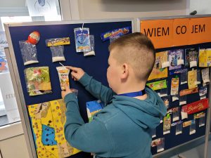 Uczeń przyczepia do tablicy opakowanie po soczku w kartoniku. Obok przyczepione są już inne produkty wraz z torebkami z cukrem.